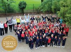 Children and staff standing in a big group in the school playground waving hands in the air to celebrate winning an award