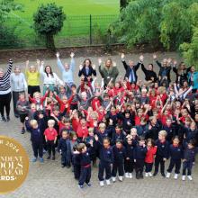 Children and staff standing in a big group in the school playground waving hands in the air to celebrate winning an award